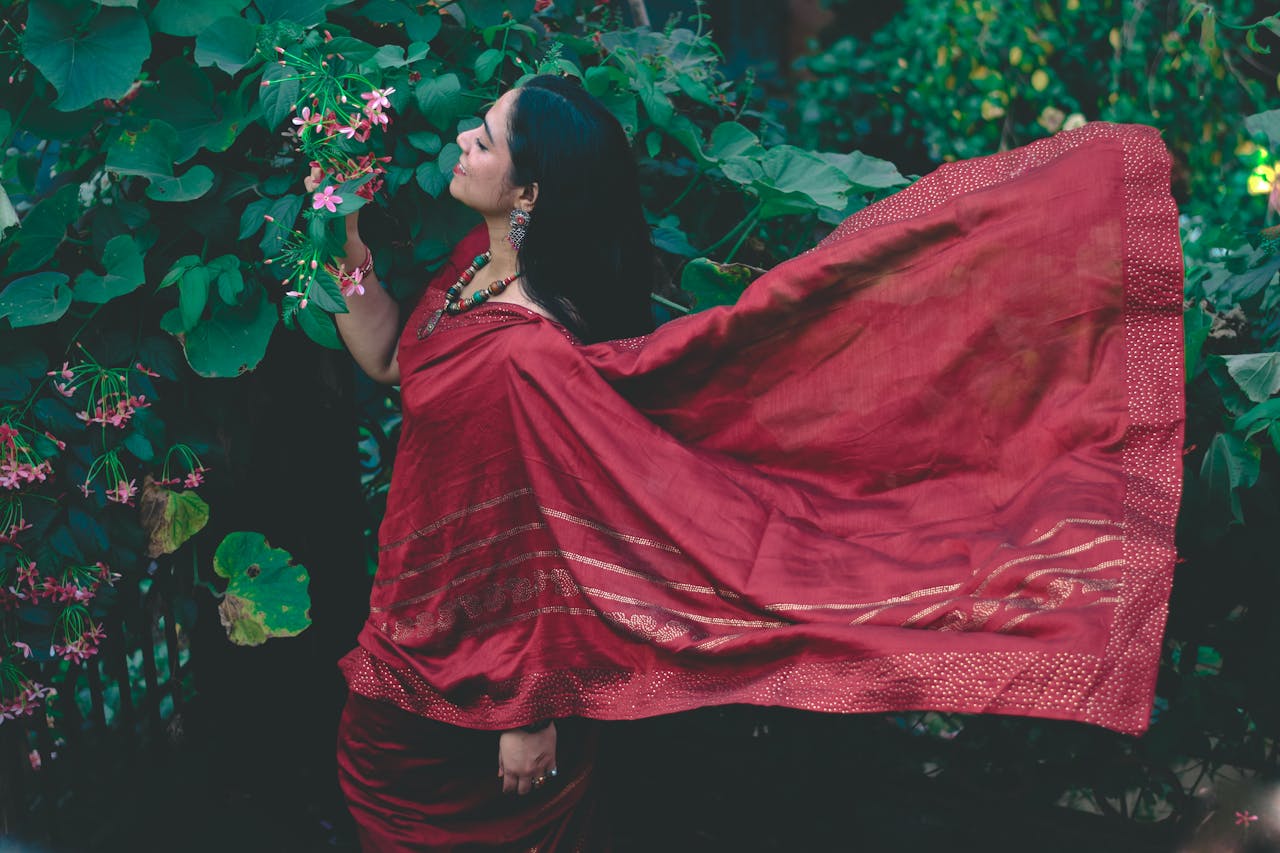 Woman in a flowing red saree stands among lush green foliage, embracing traditional Indian beauty.